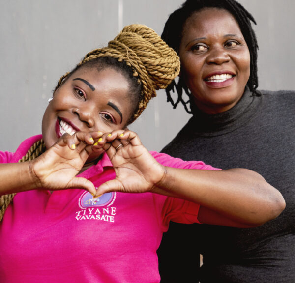 A woman in black and one in a pink shirt, the latter forming a heart with her hands